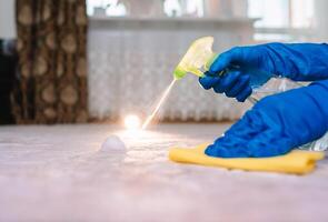 people, housework and housekeeping concept - close up of woman in rubber gloves with cloth and derergent spray cleaning carpet at home. close up of woman with cloth cleaning carpet photo