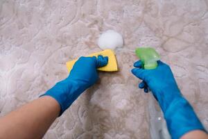 people, housework and housekeeping concept - close up of woman in rubber gloves with cloth and derergent spray cleaning carpet at home. close up of woman with cloth cleaning carpet photo