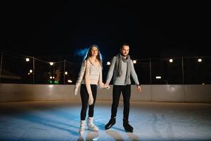 Man and woman young family Happy smiles hold hands skate on winter rink at night, with bokeh lights. photo