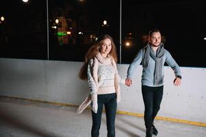 Pareja en el ciudad pista en un invierno noche. chico Ayudar bonito niña a patinar en el hielo en el oscuro noche y centelleos Encendiendo encima ellos foto