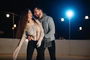 hombre y mujer joven familia contento sonrisas sostener manos patinar en invierno pista a noche, con bokeh luces. foto