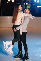 Pareja en el ciudad pista en un invierno noche. chico Ayudar bonito niña a patinar en el hielo en el oscuro noche y centelleos Encendiendo encima ellos foto