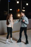 Pareja en el ciudad pista en un invierno noche. chico Ayudar bonito niña a patinar en el hielo en el oscuro noche y centelleos Encendiendo encima ellos foto