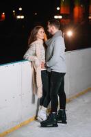 Theme ice skating rink and loving couple. meeting young, stylish people ride by hand in crowd on city skating rink lit by light bulbs and lights. Ice skating in winter for Christmas on ice arena. photo