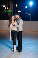 Pareja en el ciudad pista en un invierno noche. chico Ayudar bonito niña a patinar en el hielo en el oscuro noche y centelleos Encendiendo encima ellos foto