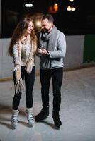 Pareja en el ciudad pista en un invierno noche. chico Ayudar bonito niña a patinar en el hielo en el oscuro noche y centelleos Encendiendo encima ellos foto