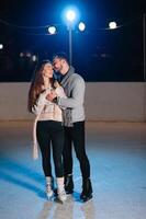 Couple on the city rink in a winter evening. Guy helping nice girl to skate on the ice in the dark night and twinkles lighting above them photo