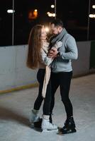 Pareja en el ciudad pista en un invierno noche. chico Ayudar bonito niña a patinar en el hielo en el oscuro noche y centelleos Encendiendo encima ellos foto