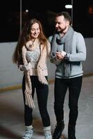 Pareja en el ciudad pista en un invierno noche. chico Ayudar bonito niña a patinar en el hielo en el oscuro noche y centelleos Encendiendo encima ellos foto