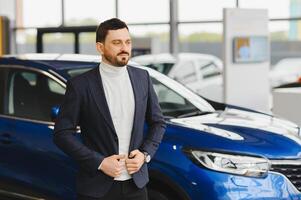 Handsome bearded buyer in casual wear in dealership, guy looks on camera while standing near car with crosed arms photo