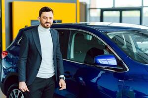 Handsome bearded buyer in casual wear in dealership, guy looks on camera while standing near car with crosed arms photo