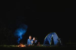 Happy romantic couple travelers resting at bonfire near glowing tourist tent under amazing night sky. The concept of active recreation and travel with a tent photo