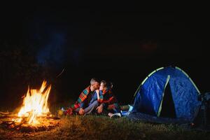 joven Pareja teniendo un descanso a hoguera junto a acampar y azul turista carpa, Bebiendo té, disfrutando noche cielo. el concepto de activo recreación y viaje con un tienda foto