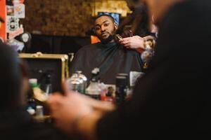 Portrait of young black man being trimmed with professional electric clipper machine in barbershop.Male beauty treatment concept. Young African guy getting new haircut in barber salon photo