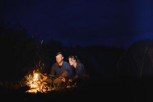 Pareja horneando salchichas en el fuego, y relajante por el hoguera en el bosque en el noche. romántico hoguera concepto. foto