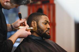 Portrait of young black man being trimmed with professional electric clipper machine in barbershop.Male beauty treatment concept. Young African guy getting new haircut in barber salon photo