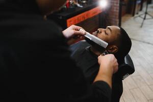 un Barbero es yendo mediante el eléctrico corte y afeitado máquina para el barba de un afroamericano brasileño chico. foto