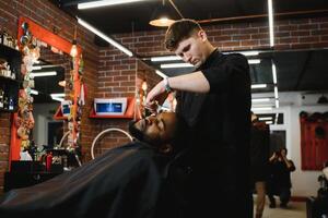 lado ver de grave hombre con elegante moderno Corte de pelo mirando adelante en Barbero tienda. mano de Barbero acuerdo Derecho maquinilla de afeitar y corte de moda rayas en cabeza de cliente. foto