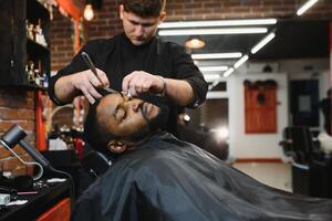 lado ver de grave hombre con elegante moderno Corte de pelo mirando adelante en Barbero tienda. mano de Barbero acuerdo Derecho maquinilla de afeitar y corte de moda rayas en cabeza de cliente. foto