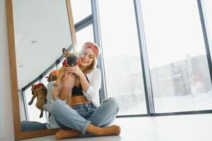 Cheerful young woman holding her big puppy with black nose and laughing. Indoor portrait of smiling girl posing with french bulldog photo