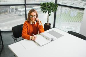 Happy smiling business woman at work talking on phone, sitting at her working place in office, copy space photo