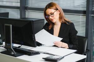 Young businesswoman sitting at workplace and reading paper in office photo