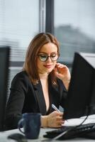 bonito, lindo, lindo, Perfecto mujer sentado a su escritorio en cuero silla en trabajo estación, vistiendo anteojos, Ropa formal, teniendo ordenador portátil y cuaderno en el mesa foto