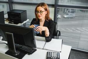 bonito, lindo, lindo, Perfecto mujer sentado a su escritorio en cuero silla en trabajo estación, vistiendo anteojos, Ropa formal, teniendo ordenador portátil y cuaderno en el mesa foto