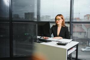 bonito, lindo, lindo, Perfecto mujer sentado a su escritorio en cuero silla en trabajo estación, vistiendo anteojos, Ropa formal, teniendo ordenador portátil y cuaderno en el mesa foto