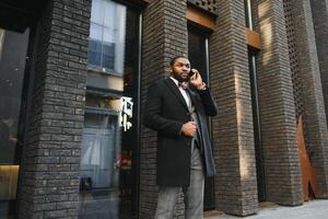 Business communications. Happy young African man in formal wear talking on the mobile phone and smiling while standing outdoors photo