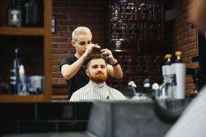 Making haircut look perfect. Young bearded man getting haircut by hairdresser while sitting in chair at barbershop. photo