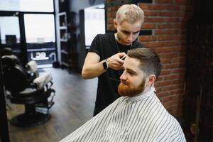 Making haircut look perfect. Young bearded man getting haircut by hairdresser while sitting in chair at barbershop. photo