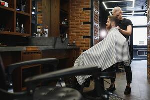 Making haircut look perfect. Young bearded man getting haircut by hairdresser while sitting in chair at barbershop photo