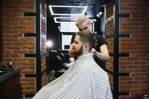 Making haircut look perfect. Young bearded man getting haircut by hairdresser while sitting in chair at barbershop. photo