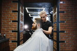 Making haircut look perfect. Young bearded man getting haircut by hairdresser while sitting in chair at barbershop. photo