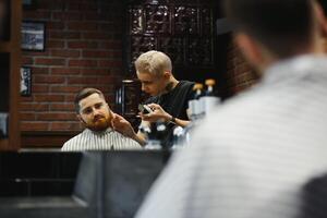 Making haircut look perfect. Young bearded man getting haircut by hairdresser while sitting in chair at barbershop. photo