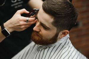 Making haircut look perfect. Young bearded man getting haircut by hairdresser while sitting in chair at barbershop. photo