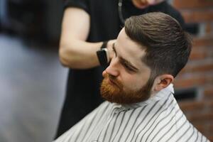 Making haircut look perfect. Young bearded man getting haircut by hairdresser while sitting in chair at barbershop. photo