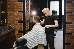 Making haircut look perfect. Young bearded man getting haircut by hairdresser while sitting in chair at barbershop. photo