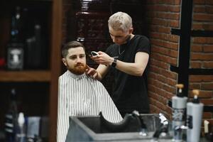 Making haircut look perfect. Young bearded man getting haircut by hairdresser while sitting in chair at barbershop. photo