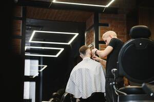 Making haircut look perfect. Young bearded man getting haircut by hairdresser while sitting in chair at barbershop. photo