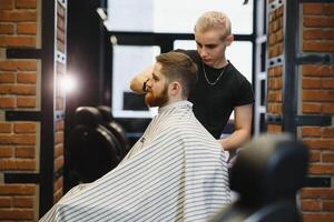 Making haircut look perfect. Young bearded man getting haircut by hairdresser while sitting in chair at barbershop. photo