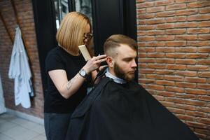 Stylish man sitting barber shop Hairstylist Hairdresser Woman cutting his hair Portrait handsome happy young bearded caucasian guy getting trendy haircut Attractive barber girl working serving client. photo