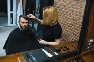 Serving client in Barbershop. Professional barber girl, female hairdresser making modern haircut for a man sitting in barber shop chair. Focus on a girl. Hairdressing, shaving, trimming, grooming. photo