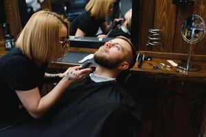 Skilled female barber cutting a beard of brunette adult guy in professional retro barbershop. photo