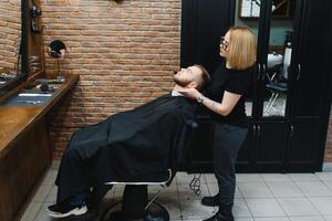 Young adult beautiful caucasian woman hairdresser cuts beard handsome man at modern barbershop Happy gay sitting chair men beauty salon. beard care. metrosexual. Female barber in black gloves serving photo