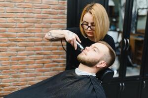 Young adult beautiful caucasian woman hairdresser cuts beard handsome man at modern barbershop Happy gay sitting chair men beauty salon. beard care. metrosexual. Female barber in black gloves serving photo
