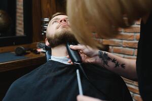 Skilled female barber cutting a beard of brunette adult guy in professional retro barbershop. photo