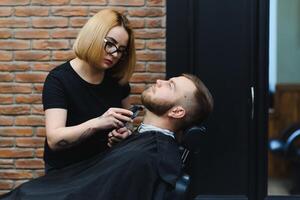 Young adult beautiful caucasian woman hairdresser cuts beard handsome man at modern barbershop Happy gay sitting chair men beauty salon. beard care. metrosexual. Female barber in black gloves serving photo