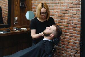 Skilled female barber cutting a beard of brunette adult guy in professional retro barbershop. photo